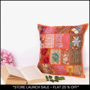 May include: A decorative patchwork cushion with an orange trim. The cushion displays a patchwork design with floral embroidery in pink, orange, and teal. An open book is placed next to the cushion.