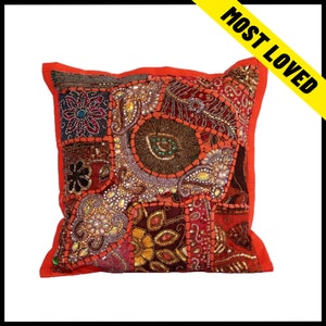 May include: Square cushion with a patchwork design. The cushion features a vibrant mix of colours including red, orange, and gold. The patchwork is embellished with sequins and beads. The text "MOST LOVED" is on a yellow banner.