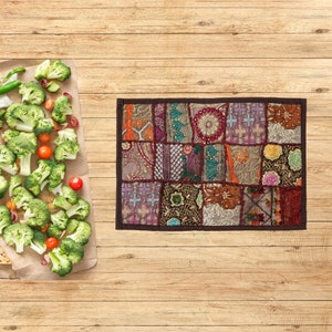 Puede incluir: Textil rectangular de patchwork con borde marrón oscuro. El textil presenta una variedad de cuadrados estampados en tonos marrón, naranja, morado y verde. Ramilletes de brócoli, tomates cherry y pimientos verdes están dispuestos sobre papel marrón.