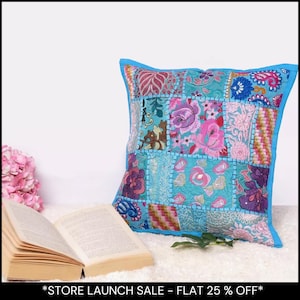 May include: Square patchwork cushion with a turquoise background and floral patterns in pink, purple, and blue. The cushion has a bright blue border. An open book and pink flowers are in the foreground. The text at the bottom says "STORE LAUNCH SALE - FLAT 25% OFF".