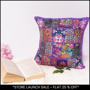 May include: A square, patchwork pillow with a purple border and a variety of embroidered designs in shades of pink, green, and gold. The pillow is resting on a white surface next to an open book and a few leaves.
