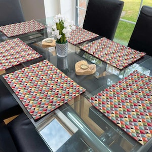 May include: Four colourful geometric patterned placemats on a glass table top. The placemats are red, yellow, green, blue, and brown.