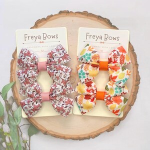 May include: Two sets of hair bows with autumn leaf patterns. The bows are made of white fabric with orange, red, and green leaves. The bows are attached to orange clips. The card reads "Freyja Bows Handmade with Love".