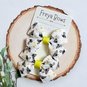 May include: Three white hair bows with a bee pattern and yellow centres are displayed on a wooden surface. The bows are attached to a card that reads "Freya Bows" with "Handmade with Love" below.