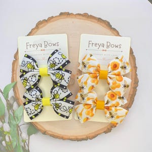 May include: Two sets of hair bows with a bee theme. One set features bees and honeycomb on a white background. The other set features bees and sunflowers on a white background. The bows are handmade and come on a card that says "Freyja Bows Handmade with Love".