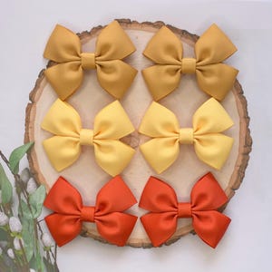 May include: Six hair bows in three colours: mustard yellow, yellow, and orange. The bows are arranged in pairs on a wooden surface.