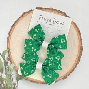 May include: Two green hair bows with white centres and a clover pattern, presented on a wooden slice. The bows are attached to a card that reads "Freya Bows, Handmade with Love."