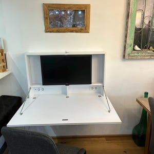 Könnte beinhalten: Ein weißer, an der Wand montierter Schreibtisch mit einer herunterklappbaren Tischplatte und einem eingebauten Monitor. Der Schreibtisch ist geschlossen und der Monitor ist sichtbar. Im Vordergrund befindet sich ein schwarzer Stuhl und im Hintergrund ein Holztisch mit einer Glasflasche und einem Stuhl.