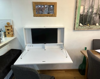 May include: A white wall-mounted desk with a fold-down top and a built-in monitor. The desk is closed, and the monitor is visible. There is a black chair in the foreground and a wooden table with a glass bottle and a chair in the background.