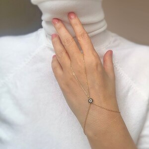May include: A gold chain bracelet with a small, round charm featuring a black and white eye design. The bracelet is worn on a person's wrist and hand, with the chain extending from the wrist to the middle finger.