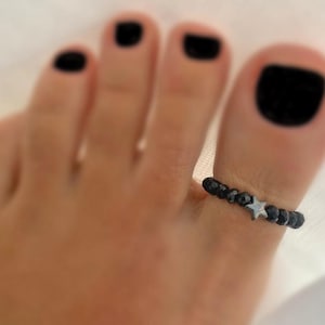 Puede incluir: Un anillo de dedo del pie con cuentas negras y un dije de estrella plateada. El anillo se lleva en un pie con esmalte de uñas negro. Una joya sencilla pero elegante.