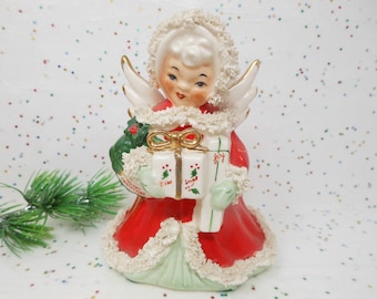 May include: A vintage ceramic Christmas angel figurine. The angel wears a red dress with white trim, holding a gift box and a green bag. The box is decorated with holly and the word "joy". The angel has white wings and a white halo.