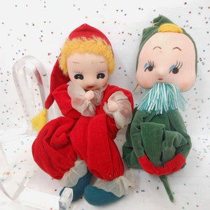 May include: Two plush dolls with painted faces and fabric clothing. One doll wears a red velvet outfit with a matching pointed hat and yellow hair. The other doll wears a green velvet outfit with a green pointed hat and a blue fringed collar.