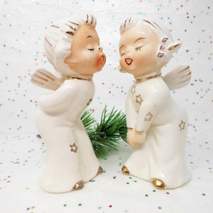 May include: Two vintage ceramic angel figurines. The angels are dressed in white robes with gold star accents and gold trim. They have closed eyes and are posed as if kissing. The wings and hair are also accented with gold.