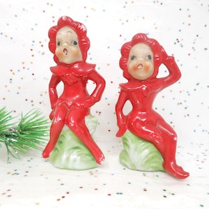May include: Two vintage ceramic figurines of seated elves in bright red outfits. The elves have pale skin, rosy cheeks, and are perched on green and white bases. The background is white with colorful confetti.