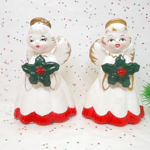 May include: Two vintage ceramic angel figurines. Each angel is dressed in a white dress with a red scalloped hem and holds a green star-shaped ornament with red berries. They have gold halos and wings.