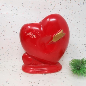 May include: Red heart-shaped ceramic vase with a gold arrow design. The vase is sitting on a white surface with glitter.