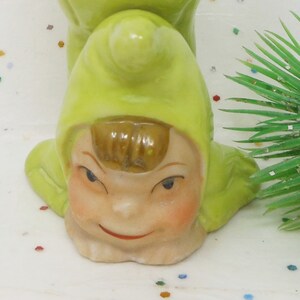 May include: A vintage ceramic figurine of a smiling elf with a green hat and face. The elf has rosy cheeks, brown hair, and a red smile. The figurine is on a white surface with colorful confetti and a green decorative branch.