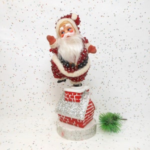 May include: A vintage Christmas decoration featuring Santa Claus atop a red brick chimney with a silver glitter roof. Santa is dressed in a red suit with white trim and a long white beard. The chimney sits on a wooden base.