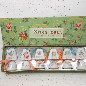 May include: A set of twelve Christmas bells in a decorative box. The bells are white with various holiday-themed illustrations and greetings. The box is green with a festive pattern and the text "Xmas Bell Set of 12".