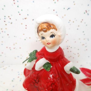 May include: A ceramic figurine of a child wearing a red dress with white trim and a white bonnet. The figure has brown hair, painted facial features, and holds a green sprig of holly with red berries in one hand, finished with a glossy glaze.