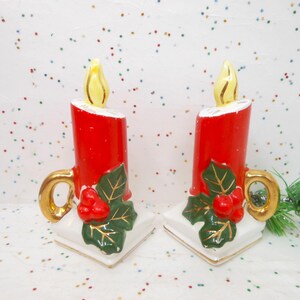May include: Pair of ceramic candle holders. Each candle is red with a yellow flame, gold handle, and white base. Green leaves and red berries decorate the front. Festive holiday decor.
