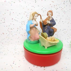 May include: A Christmas nativity scene figurine set. The set features Mary, Joseph, and baby Jesus in a manger. The figurines are on a green base atop a red, round box. The figures are painted in muted colors.