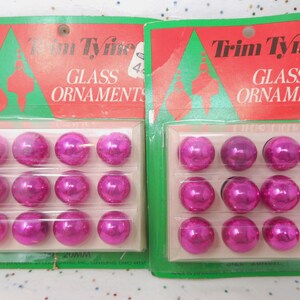 May include: Two packages of Trim Tyne glass ornaments. Each package contains twelve shiny, magenta-colored, 20mm glass ball ornaments. The packaging has a red and green design with the words "Glass Ornaments".