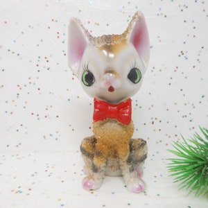 May include: A ceramic figurine of a cat wearing a red bow tie. The cat has brown and white fur and large green eyes. The figurine is sitting on a white background with colorful confetti.