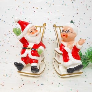May include: A pair of ceramic Christmas figurines depicting Santa Claus and Mrs. Claus in rocking chairs. Santa wears a red suit with a black belt and green gloves. Mrs. Claus wears a red coat and glasses. The chairs are white with gold accents.