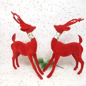 May include: Two red velvet reindeer figurines with silver and green polka dot collars. The reindeer have long, slender legs and antlers. A sprig of greenery is placed between them. Festive holiday decor.