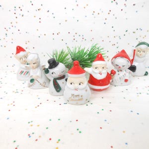 May include: A collection of vintage Christmas figurines, including Santa Claus, snowmen, and angels. The figurines are white with red, green, and black accents. The Santa figurine has the words "Merry Christmas" on it. The figurines are on a white surface with colorful confetti.