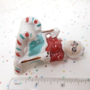K&ouml;nnte beinhalten: Kleine Santa Claus-Figur aus Keramik, die auf einem Zuckerstangenstuhl sitzt. Die Santa-Figur hat einen roten Mantel, einen wei&szlig;en Bart und eine Brille. Der Zuckerstangenstuhl ist wei&szlig; mit roten und t&uuml;rkisfarbenen Streifen. Ca. 5 cm lang.