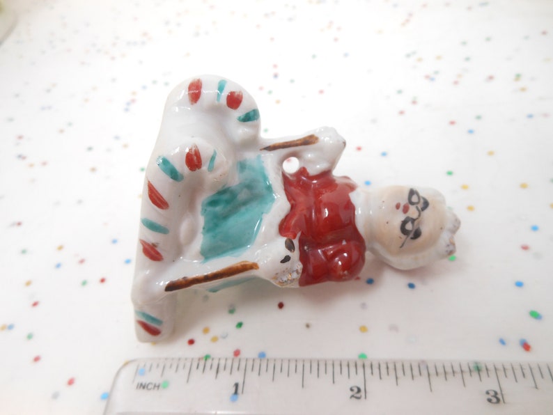 K&ouml;nnte beinhalten: Kleine Santa Claus-Figur aus Keramik, die auf einem Zuckerstangenstuhl sitzt. Die Santa-Figur hat einen roten Mantel, einen wei&szlig;en Bart und eine Brille. Der Zuckerstangenstuhl ist wei&szlig; mit roten und t&uuml;rkisfarbenen Streifen. Ca. 5 cm lang.