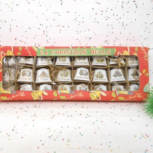 May include: A box of 24 Christmas bells. The bells are white with gold accents and feature various holiday-themed designs. The box is red with a green banner that reads "24 CHRISTMAS BELLS".