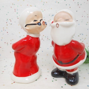 May include: A pair of ceramic figurines depicting Mrs. Claus and Santa Claus leaning toward each other. Mrs. Claus wears a red dress with white trim and black glasses. Santa Claus wears a red suit with a white beard, black belt, and black boots.