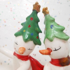 May include: A pair of ceramic snowman figurines. Each snowman has a white body, carrot nose, and a green Christmas tree hat with colorful ornaments. They wear red scarves and have closed eyes, suggesting a festive theme.