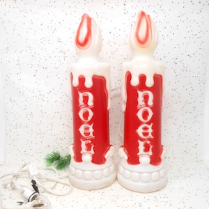 May include: Two red and white Christmas candle lights with the word "NOEL" in white. The candles have a dripping wax design and a flame-shaped top. The base is white with a decorative design. The lights are plugged in.
