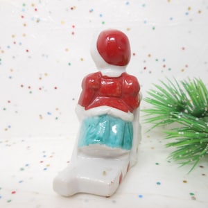 K&ouml;nnte beinhalten: Kleine Keramikfigur einer sitzenden Figur, die eine rote Jacke und einen Hut sowie einen t&uuml;rkisfarbenen Rock tr&auml;gt. Die Figur steht auf einem wei&szlig;en Sockel und ist wahrscheinlich eine Weihnachtsdekoration.