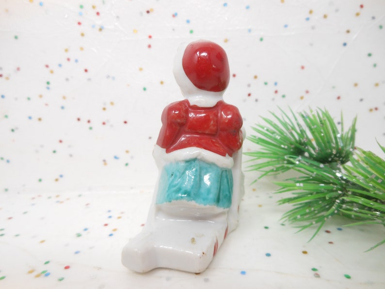 K&ouml;nnte beinhalten: Kleine Keramikfigur einer sitzenden Figur, die eine rote Jacke und einen Hut sowie einen t&uuml;rkisfarbenen Rock tr&auml;gt. Die Figur steht auf einem wei&szlig;en Sockel und ist wahrscheinlich eine Weihnachtsdekoration.