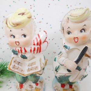 May include: Two vintage ceramic Christmas carolers. One holds sheet music, the other a violin. Both figures have blonde hair, rosy cheeks, and are dressed in white and green outfits with red accents. The figurines are on a snowy base.