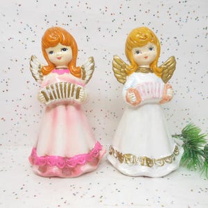 May include: Two ceramic angel figurines. One angel wears a pink dress and the other a white dress. Both have golden wings and hold accordions. The angels have orange and blonde hair, respectively. The figurines are decorative and likely for holiday display.