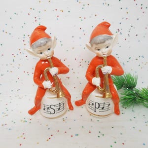 May include: Two ceramic elf figurines in orange suits and hats, playing gold trumpets. They sit atop white bases with musical notes and the letters "S" and "P". A festive holiday decoration.