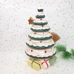 May include: A ceramic Christmas tree-shaped container with a white base, green trim, and red accents. The tree has gold star and bell-shaped labels marked "P" and "S". The base features yellow and green gift boxes.