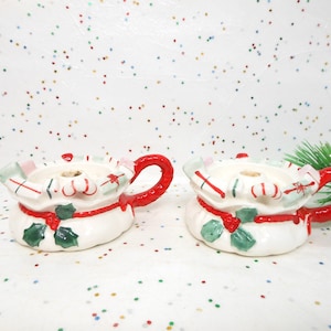 May include: Two white ceramic Christmas candle holders shaped like gift bags, with red handles and accents. Each holder features green holly leaves, red ribbons, and candy cane designs. Festive holiday decor.