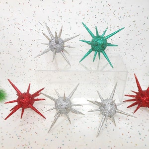 May include: A collection of spiky, star-shaped ornaments in various colors, including silver, red, and green. The ornaments are covered in glitter and are arranged on a clear surface, suggesting a festive or holiday theme.