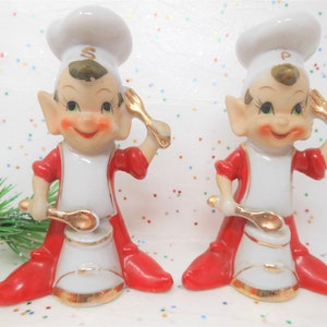 May include: A pair of vintage ceramic salt and pepper shakers. Each shaker is a smiling chef elf wearing a white chef's hat, red outfit, and holding a golden spoon. The salt shaker has an "S" on the hat, and the pepper shaker has a "P".