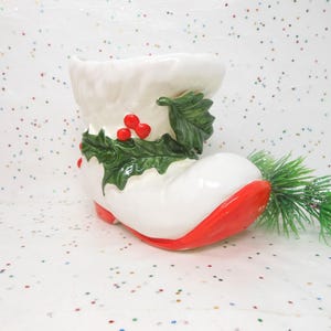 May include: A white ceramic Christmas boot-shaped planter with a red sole and accents. The boot is decorated with green holly leaves and red berries. A small sprig of green pine is next to the boot.