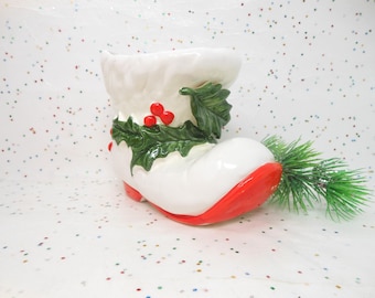 May include: A white ceramic Christmas boot-shaped planter with a red sole and accents. The boot is decorated with green holly leaves and red berries. A small sprig of green pine is next to the boot.
