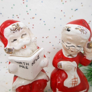 May include: A pair of vintage ceramic Christmas figurines. One is Santa Claus reading a newspaper with "North Pole News" printed on it. The other is Mrs. Claus holding a sack. Both are wearing red and white outfits.